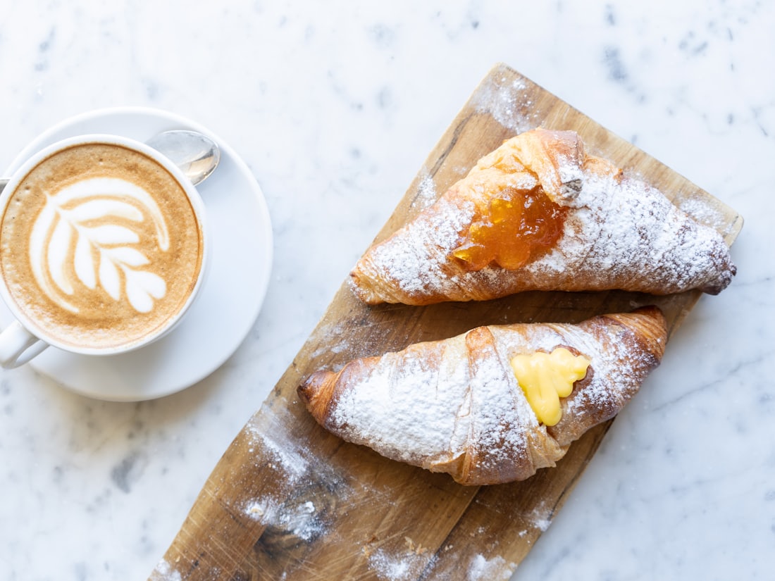 Cappuccino mit Milchschaummuster daneben zwei gefüllte Plundergebäcke auf Holzbrett